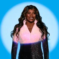 A woman, Marcia Hines, stands in a spotlight wearing a pink glitter top with long brown wavy hair flowing over her shoulders